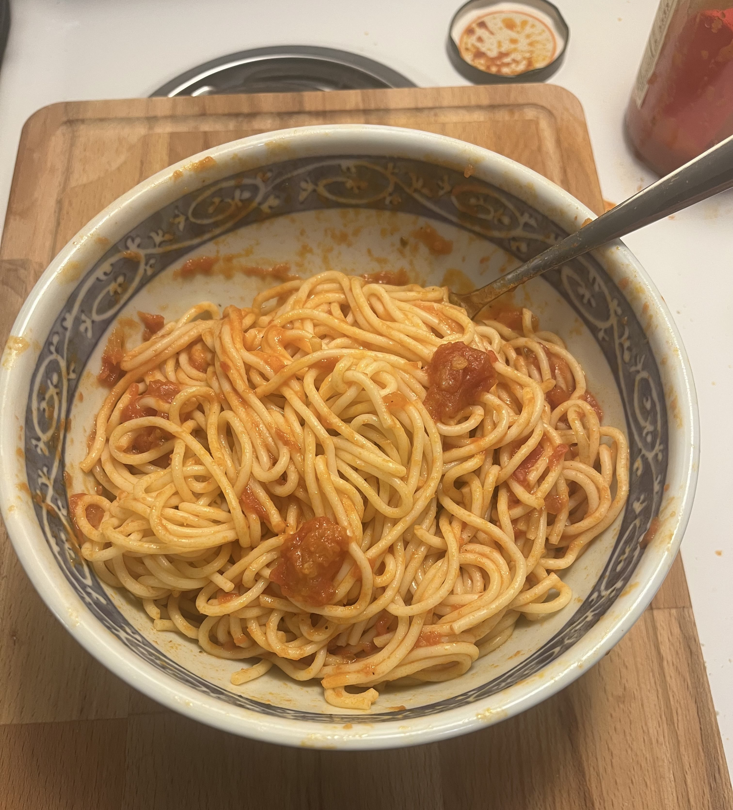 pasta in a bowl with a fork sticking out of it. bowl is sitting on top of a cutting board, which is on top of a stove. in the background, there is a jar of marinara sauce with a red silicon spatula-type cooking tool resting inside. this jar is also on top of the stove.